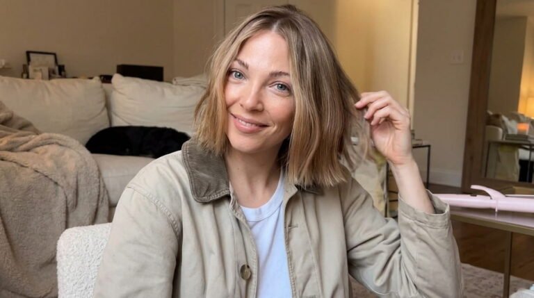 Smiling woman with a shoulder-length blonde bob and honey highlights, touching her hair, wearing a beige corduroy-collar jacket over a white tee in a cozy living room
