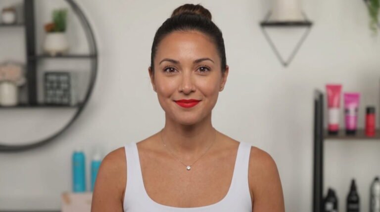 Smiling woman with dark hair in a high bun, red lipstick and a thin pendant necklace, wearing a white tank top in front of shelves with hair care products